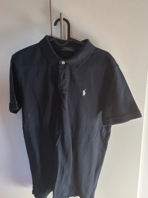 Marinblå pikétröja från Polo Ralph Lauren - Klassisk marinblå pikétröja från Polo Ralph Lauren med vit broderad logga på bröstet. Tröjan har krage, knappar framtill och korta ärmar. Perfekt till jeans eller shorts för en clean och tidlös look.