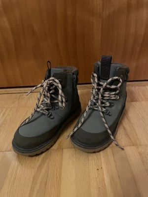 Grå, gröna fodrade kängor med snörning - Snygga grå kängor med beige och svarta snören, grov sula och dragkedja på sidan. Insidan är fodrad med mjukt, brunt foder för extra värme. Perfekta för kalla dagar och har en modern, robust look.