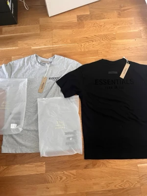 Essentials Fear of God 2 t-shirts - Två snygga Essentials Fear of God t-shirts, en i ljusgrått och en i svart. Båda har klassisk rund hals, korta ärmar och tryck med logga fram och bak. Perfekta till streetwear-looken och tillverkade i mjuk bomull för maximal komfort.