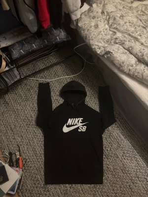 Svart Nike SB hoodie med logga - Svart hoodie från Nike SB med stor vit logga på bröstet. Klassisk huva och lång ärm, perfekt för en avslappnad streetwear-look. Tillverkad i mjukt material som känns skönt mot huden. Enkel och stilren design som passar till det mesta.