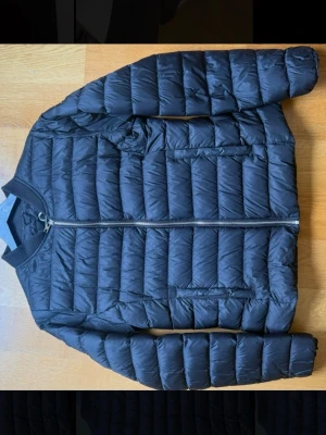 Svart lättvikts pufferjacka - Snygg svart pufferjacka med snygg design och dragkedja framtill. Jackan har två sidofickor med dragkedja och ribbad krage. Tillverkad i ett lätt och smidigt material som håller värmen utan att kännas klumpig. Perfekt för dig som vill ha en stilren och enkel look.