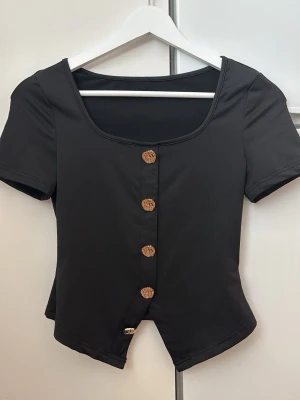 Svart topp med träknappar - Snygg svart topp med korta ärmar och dekorativa träknappar framtill. Toppen har en något croppad passform och en bred, rundad urringning. Perfekt att styla med jeans eller kjol för en trendig look.
