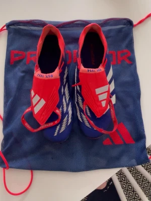 Adidas Predator blå/röda fotbollsskor - Snygga Adidas Predator fotbollsskor i blått och rött med vita detaljer. Skorna har en strumpliknande passform och tydliga Predator-loggor. Ovandelen är i syntetmaterial och sulan har dobbar för bra grepp på planen. Perfekta för dig som vill sticka ut på matchen!
