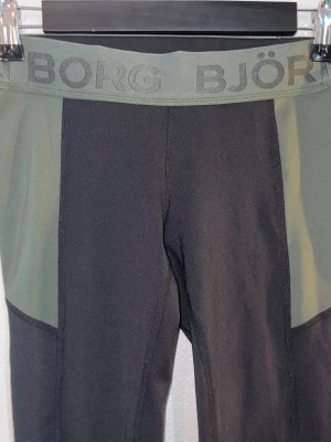 Svarta träningstights från Björn Borg - Snygga svarta träningstights från Björn Borg med mörkgröna paneler på sidorna och bred resår i midjan med logga. Tillverkade i stretchigt funktionsmaterial som andas, perfekta för gym eller löpning.