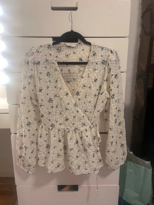 Vit blommig omlottblus från Pigalle - Söt vit omlottblus från Pigalle med blått blommönster och struktur i tyget. Blusen har långa ärmar med resår vid ärmslut och knytband i midjan för en feminin siluett. Perfekt till jeans eller kjol för en fräsch look.
