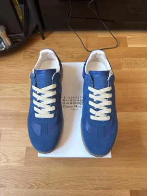 Blå sneakers Maison Margiela - Snygga blå sneakers från Maison Margiela med vita skosnören och klassisk siluett. Ovandelen är i mocka och textil med detaljer i läder. Sitter på en vit sula och har en stilren look som passar till det mesta.