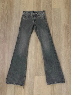 Grå bootcut low waist jeans - Ett par grå lowwaist bootcut jeans med lätt tvättad look och slitna detaljer vid bensluten. Två små hål längst ned vid benen, men går enkelt att fixa med 1cm uppsyning (Jag kan fixa vid +49kr) Jeansen har fyra fickor och dragkedja. Perfekta för en avslappnad och trendig stil. 