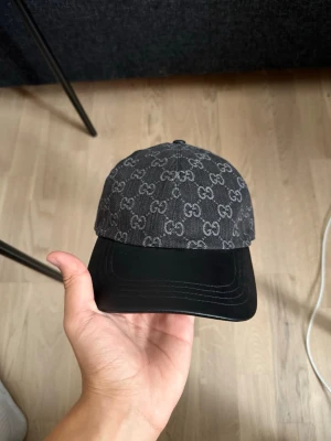 Svart keps med GG-mönster från Gucci - Snygg svart keps från Gucci med det klassiska GG-mönstret över hela ovandelen. Skärmen är i svart läderlook och resten av kepsen är i tyg. Perfekt accessoar för att lyfta din outfit och ge en exklusiv touch.