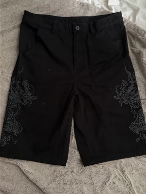 Svarta jorts med drakmönster - Säljer ett par svarta jorts med snyggt drakmönster på båda benen. Jortsen har rak passform, bälteshällor och knappstängning framtill. Perfekta för dig som vill sticka ut med coola detaljer och streetwear-vibe. Märket är Ed Hardy men köpta på Urban Oufitters.