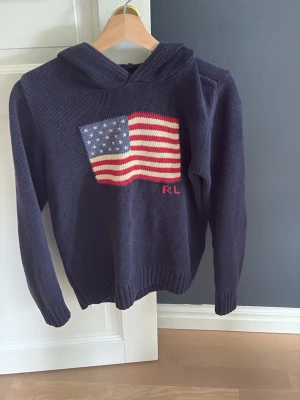 Stickad Ralph lauren tröja  - Snygg marinblå stickad hoodie med stor amerikansk flagga på bröstet och RL-broderi. Tröjan har huva, långa ärmar och ribbade muddar. Perfekt för dig som gillar klassisk och avslappnad stil.