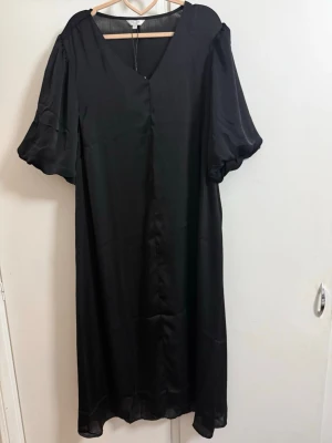 Svart långklänning med puffärm - Svart långklänning med v-ringning och korta puffärmar. Klänningen har en lös passform och är tillverkad i ett glansigt, mjukt material som ger en elegant känsla. Perfekt för dig som gillar stilrena och klassiska plagg.