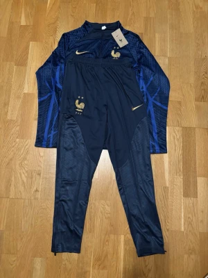 Frankrike träningsset Nike blå/guld - Snyggt träningsset från Nike i mörkblått med gulddetaljer och Frankrikes landslagsemblem. Tröjan har halv dragkedja och grafiskt mönster i blått, byxorna har smal passform och samma emblem. Perfekt för fotbollsträning eller chill.