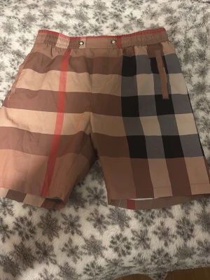 Rutig beige och brun shorts - Snygga shorts med klassiskt rutigt mönster i beige, brunt, svart och rött. Shortsen har elastisk midja med knappar och snörning samt fickor på sidorna. Perfekta för sommardagar och har en avslappnad passform.