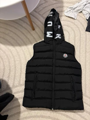 Svart dunväst med huva från Moncler - Snygg svart dunväst från Moncler med huva och dragkedja framtill. Västen har två fickor med dragkedja och Moncler-logga på bröstet. Huva med tryckt text i vitt ger en cool detalj. Perfekt för lager-på-lager och streetstyle.