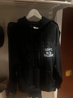Svart hoodie med tryck 'DÉPT. de la GALERIE' - Säljer en svart hoodie med huva och ficka framtill. På bröstet står det 'DÉPT. de la GALERIE' i vitt tryck och större text i svart bakom. Hoodien har dragsko i huvan och en avslappnad passform, perfekt för en chill och trendig look. Stl M passar L