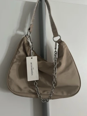 Beige axelremsväska från NLY Accessories - Stilren beige axelremsväska från NLY Accessories i mjukt material. Väskan har en bred axelrem och en dekorativ silverfärgad kedja framtill. Perfekt för att lyfta din outfit med en trendig touch. Stängs med dragkedja och har en modern, rundad form.