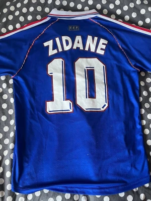 Frankrike Zidane tröja VM 98 Adidas - Klassisk blå fotbollströja från Adidas, Frankrikes landslag, med Zidane och nummer 10 på ryggen. Tröjan har vita och röda detaljer, broderad tupp-logga och FIFA World Cup France 98-tryck på bröstet. Tillverkad i lätt och ventilerande material.