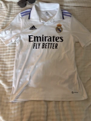 Real Madrid matchtröja Adidas vit - Snygg vit Real Madrid matchtröja från Adidas med klubbmärke och sponsortryck på bröstet. Tröjan har krage, korta ärmar och lila detaljer på axlarna. Tillverkad i ett lätt och ventilerande material, perfekt för fotboll eller träning.