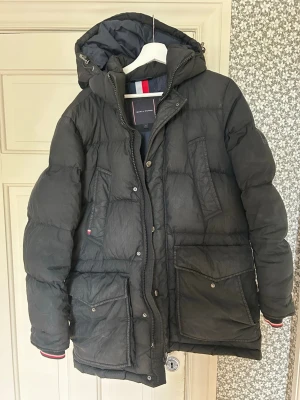 Svart dunjacka från Tommy Hilfiger - Snygg svart dunjacka i stl S (passar M) från Tommy Hilfiger med huva och dragkedja. Jackan har stora fickor med lock och tryckknappar, samt diskreta Hilfiger-detaljer. Perfekt för kalla vinterdagar tack vare det tjocka dunfodret och den höga kragen.