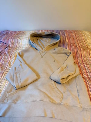 Beige hoodie från Polo Ralph Lauren - Snygg beige hoodie från Polo Ralph Lauren med klassisk huva och känguruficka framtill. Tillverkad i mjukt bomullsmaterial som känns skönt mot huden. Diskret logga broderad på bröstet och ribbade muddar vid ärmar och nederkant.