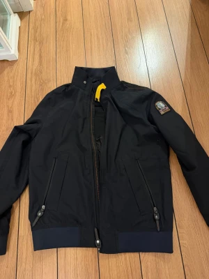 Svart vindjacka från Parajumpers - Snygg svart vindjacka från Parajumpers med hög krage, två dragkedjeförsedda fickor och ikonisk patch på ärmen. Jackan har en gul detalj vid halsen och muddar vid ärmslut och nederkant. Perfekt för blåsiga dagar och streetwear-stil.