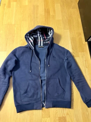 Blå hoodie med dragkedja och huva från burberry - Snygg blå hoodie med dragkedja och huva med randigt foder. Tröjan har två stora fickor framtill och ribbade muddar vid ärmar och nederkant. Broderad logga på bröstet och justerbara snören i huvan. Perfekt för en avslappnad stil. Passformen är perfekt, passar både L och M