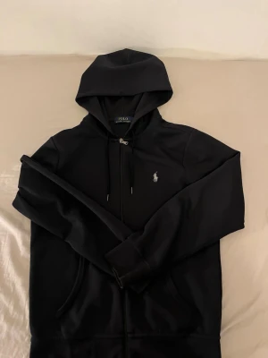 Svart hoodie från Polo Ralph Lauren - Snygg svart hoodie från Polo Ralph Lauren med dragkedja framtill och klassisk huva med dragsko. Ikoniska loggan broderad på bröstet. Tillverkad i mjukt material som känns skönt mot huden. Perfekt för en avslappnad och stilren look.