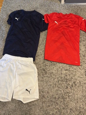 Puma fotbollskläder set - Säljer ett set med två Puma fotbollströjor, en marinblå och en röd, samt ett par vita shorts. Alla plagg har Puma-loggan tryckt på bröstet eller benet och är tillverkade i ett lätt, snabbtorkande material perfekt för träning eller match.