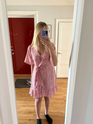 Rosa blommig omlottklänning - Söt kort rosa omlottklänning med blommigt mönster och volangkant nertill. Klänningen har v-ringning, knyt i midjan och korta ärmar. Perfekt för sommarens alla tillfällen.