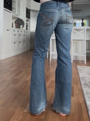 Blå bootcut jeans med broderi - Säljer ett par blå bootcut jeans med låg midja och snygga broderade detaljer på bakfickorna. Midjemått: 37 och innerbenslängd: 79