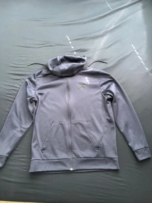 Mörkgrå hoodie från Under Armour - Snygg mörkgrå hoodie från Under Armour med dragkedja framtill och två stora fickor. Tillverkad i ett mjukt och stretchigt material som känns sportigt. Har en huva med dragsko och diskret logga på bröstet. Perfekt för chill eller träning.