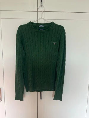 Grön kabelstickad tröja från GANT - Mysig grön kabelstickad tröja från GANT med rund halsringning och diskret logga broderad på bröstet. Tröjan har ribbade muddar vid ärmslut och nederkant. Perfekt för lager på lager och en klassisk look.
