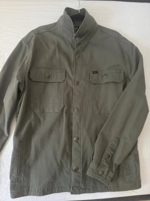 Grön overshirt från Lee - Stilren grön overshirt från Lee med två stora bröstfickor med lock och knapp, samt knappstängning framtill. Tillverkad i ett slitstarkt bomullstyg och har klassisk krage och justerbara manschetter. Perfekt lager på lager-plagg för en avslappnad look.
