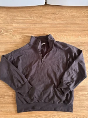 Mörkbrun halvzip tröja - Mörkbrun tröja med halv dragkedja och hög krage. Tröjan har långa ärmar, ribbade muddar och nederkant. Perfekt för lager på lager och enkel att matcha med olika outfits.