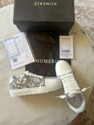 Gråvita sneakers från Numeris - Snygga sneakers från Numeris i grått och vitt med mönstrad textil på sidorna och vita detaljer. Skorna har tjock vit sula, vita skosnören och en låg profil. Perfekta för dig som vill ha en modern och stilren look.