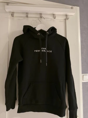 Svart hoodie Peak Performance - Säljer en svart hoodie från Peak Performance med vit logga på bröstet. Hoodien har huva med snören och en stor magficka. Tillverkad i mjukt material som känns skönt mot huden. Perfekt för en avslappnad och sportig stil.