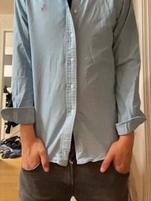 Ljusblå skjorta från Polo Ralph Lauren - Klassisk ljusblå skjorta från Polo Ralph Lauren med button-down krage och lång ärm. Skjortan har broderad logga på bröstet och vita knappar. Tillverkad i bomull för en bekväm känsla och snygg passform.
