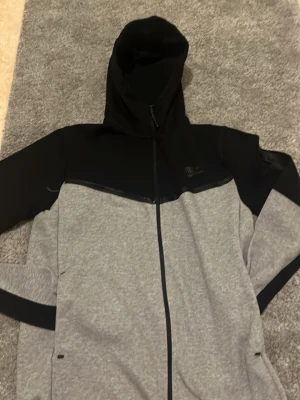 Nike grå och svart zip hoodie - Snygg hoodie från Nike i grått och svart med dragkedja framtill. Hoodien har en stor huva och diskret Nike-logga på bröstet. Tillverkad i mjukt material, perfekt för en sportig och avslappnad stil. Fickor på sidorna och svarta detaljer längs ärmarna.