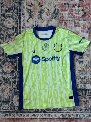 FC Barcelona matchtröja Lamine Yamal - Snygg FC Barcelona matchtröja i neongult med blå detaljer och tryck. Tröjan har Lamine Yamal och nummer 19 på ryggen, samt sponsorloggor från Spotify och UNHCR. Tillverkad i lätt och ventilerande material, perfekt för fotboll eller träning.