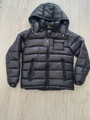 Svart pufferjacka från Maggiore - Snygg svart pufferjacka från Maggiore med huva och quiltad design. Jackan har två dragkedjeförsedda fickor framtill och en bröstficka med knapp. Perfekt för kalla vinterdagar tack vare det vadderade materialet och den höga kragen.