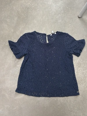 Mörkblå spetsblus med kort ärm - Snygg mörkblå blus i spets med korta ärmar och rund halsringning. Blusen har ett litet sprund bak i nacken och är tillverkad i ett lätt och luftigt material. Perfekt för dig som gillar feminina detaljer och en stilren look. Priset kan diskuteras!