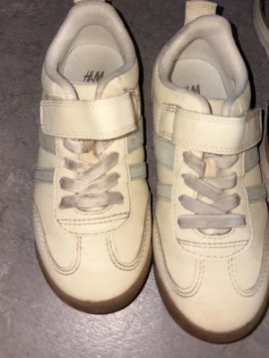 Vita sneakers med kardborre från H&M - Snygga vita sneakers från H&M med kardborreband och gråa detaljer. Skorna har rund tå, vita skosnören och beige sula. Tillverkade i syntetmaterial och textil för en bekväm känsla. Perfekta för en avslappnad och trendig stil.