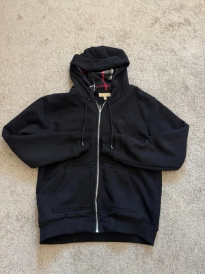 Svart hoodie från burberry - Säljer en svart hoodie med dragkedja och huva. Insidan av huvan har ett rutigt mönster i svart, vitt och rött. Hoodien har fickor framtill och ribbade muddar vid ärmslut och nederkant. Perfekt för en avslappnad och stilren look.