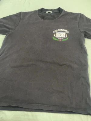 Svart t-shirt från Casablanca - Svart t-shirt från Casablanca med broderad logga och motiv på bröstet. Trycket visar ett klassiskt tempel med texten 'Equipment Sportif' och gröna detaljer. T-shirten har rund hals och korta ärmar, perfekt för en avslappnad och trendig stil.