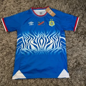 Blå Umbro fotbollströja ECOF - Snygg blå fotbollströja från Umbro med vit zebramönstrad design på framsidan. Tröjan har röda och vita detaljer vid ärmslut och krage, samt ECOF-emblem och flagga på bröstet. Perfekt för match eller träning.