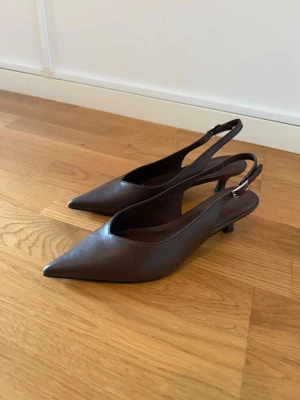 Bruna slingback pumps med låg klack - Stilrena bruna pumps med spetsig tå och slingback-rem. Skorna har en låg klack och justerbart spänne vid hälen. Tillverkade i ett slätt material som ger en elegant look. Perfekta för dig som gillar klassisk och tidlös stil. Endast använda en gång. Nypris 340kr