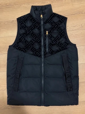 Svart dunväst från Glorious Gangsta - Snygg svart dunväst från Glorious Gangsta med hög krage och guldfärgade dragkedjor. Överdel i sammet med mönstrad design och två fickor framtill. Perfekt för dig som vill ha en streetinspirerad look med exklusiva detaljer.