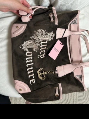 Juicy Couture brun och rosa handväska - Juicy Couture handväska i brun mocka med ljusrosa detaljer och handtag. Väskan har dekorativa broderier, metallkedja med krona och logga framtill. Rymlig modell med innerfack och snygga kontrastdetaljer. Perfekt för dig som gillar en trendig och klassisk look. Köpt från usa i mycket god skick 