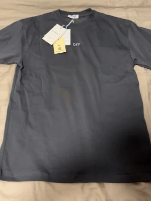 Mörkgrå Off-White t-shirt - Snygg mörkgrå t-shirt från Off-White med diskret 'OFF' tryck fram och stort ikoniskt pilar-tryck på ryggen. Klassisk passform och rund halsringning. Tillverkad i mjuk bomull, perfekt för en clean streetwear-look.