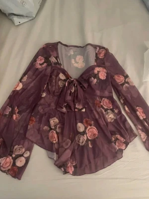 Lila blommig meshblus med rosett - Snygg lila meshblus med rosa och röda rosor, v-ringning och stor rosett framtill. Blusen har långa, vida ärmar och är tillverkad i ett tunt, transparent material som ger en luftig känsla. 